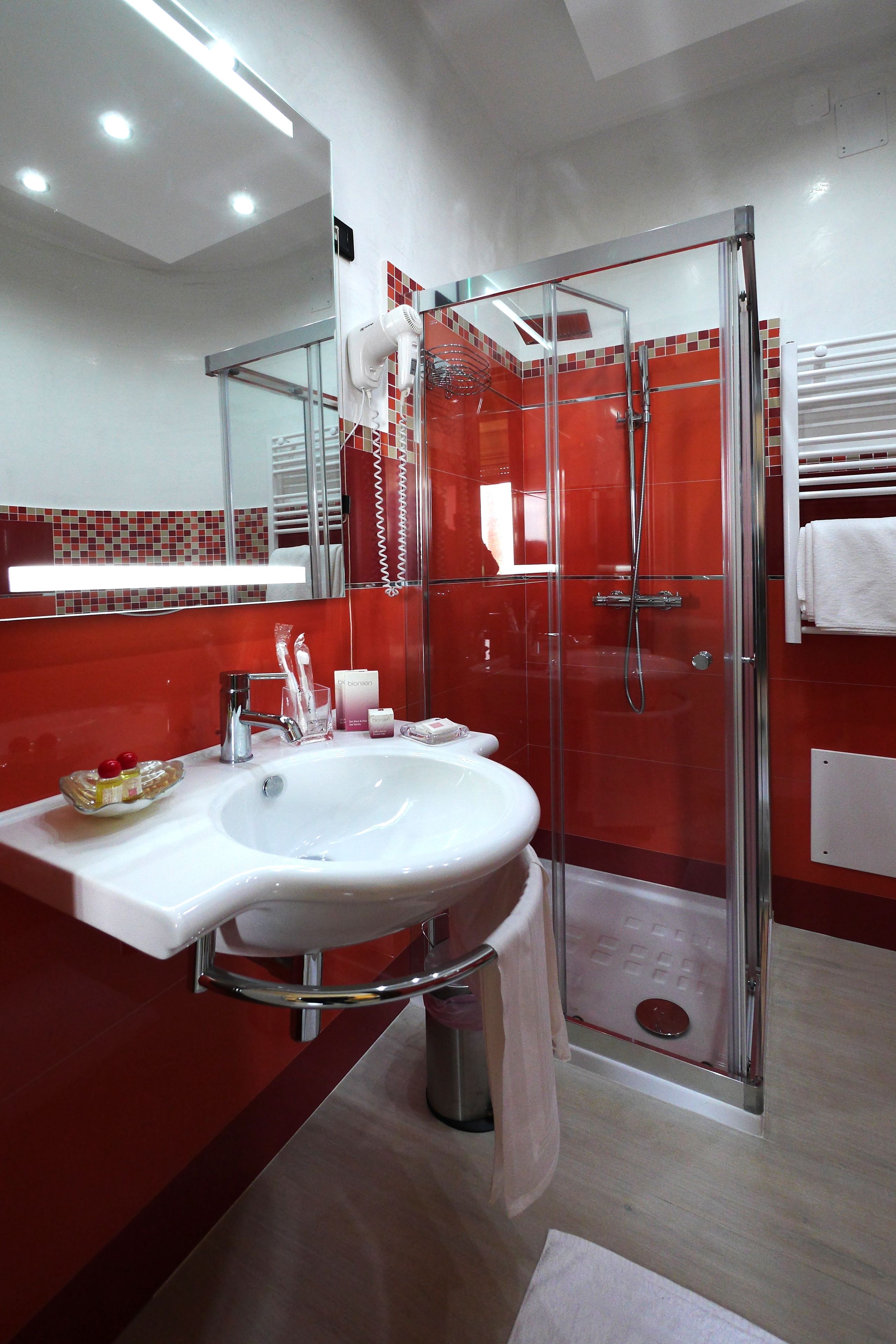 Un baño moderno con cabina de ducha de vidrio, un lavamanos blanco y una pared revestida de azulejos rojos.