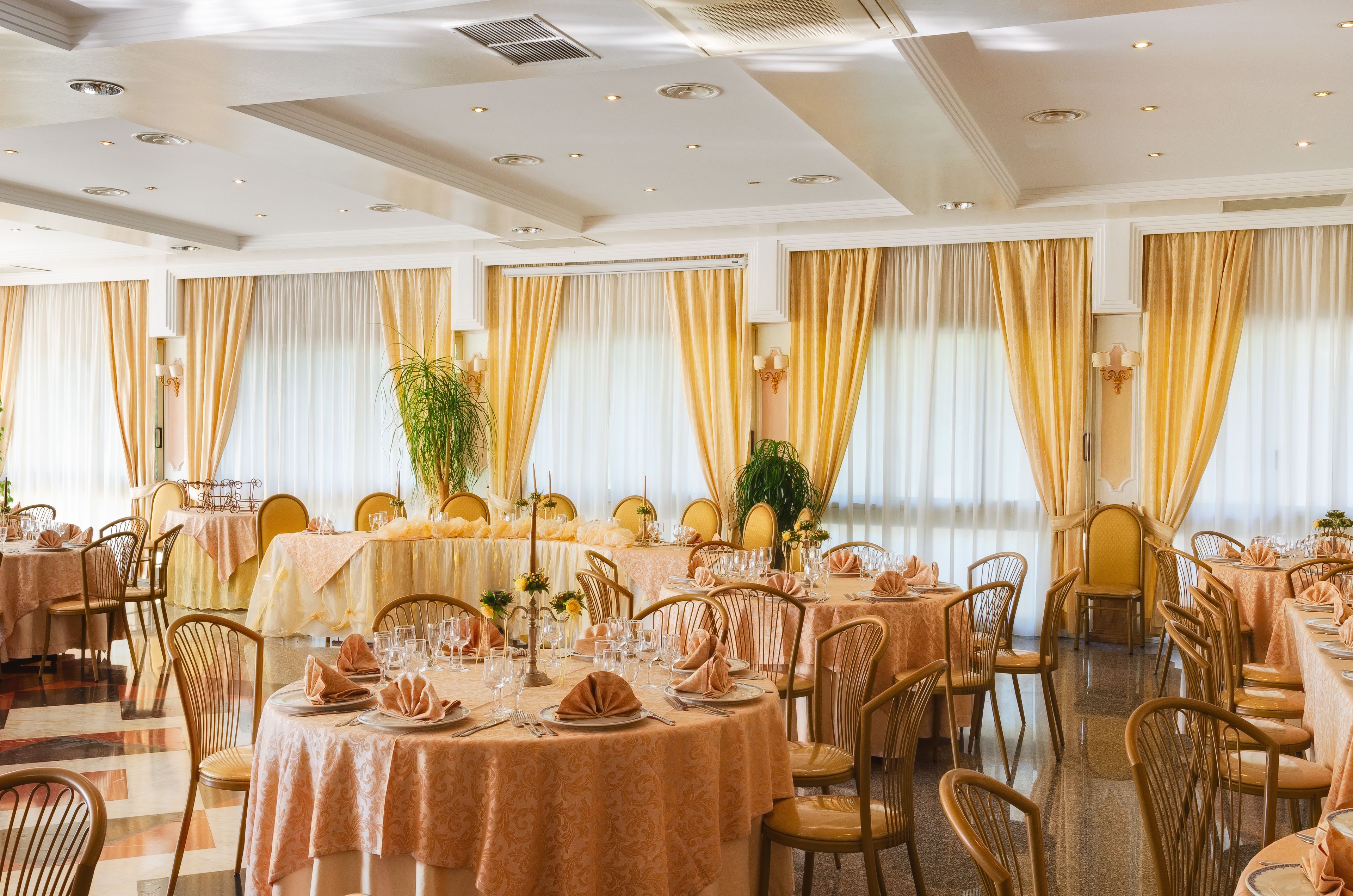 A banquet hall with round tables, golden curtains, and chandeliers.