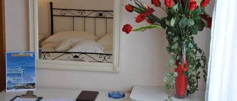 Ein Hotelzimmer mit Bett, Schreibtisch mit Spiegel, einer Vase mit roten Blumen und einem Infobrett.