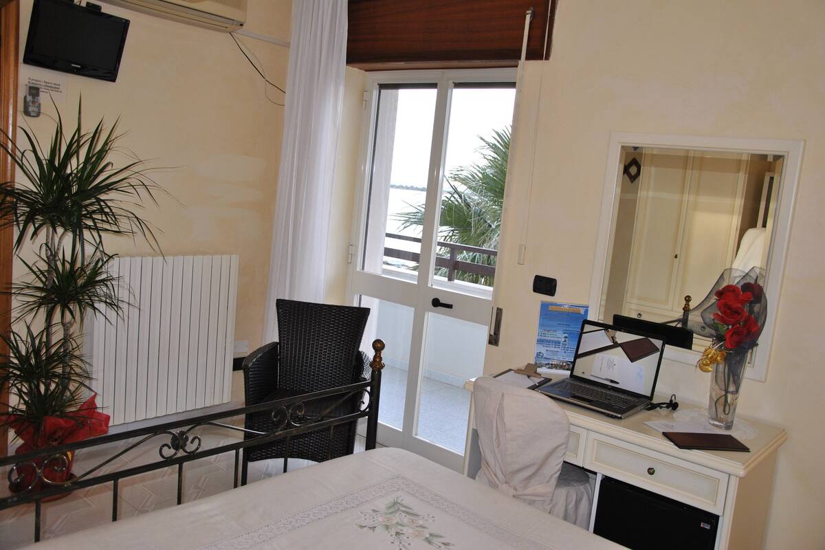 Une chambre d’hôtel comprenant un lit, un bureau avec un ordinateur portable, une chaise, une plante en pot, une fenêtre donnant sur des palmiers et un téléviseur fixé au mur.
