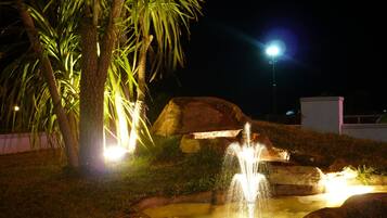 Nachtszenerie im Freien mit einem Springbrunnen, beleuchtet durch künstliches Licht, umgeben von Felsen und Palmen.