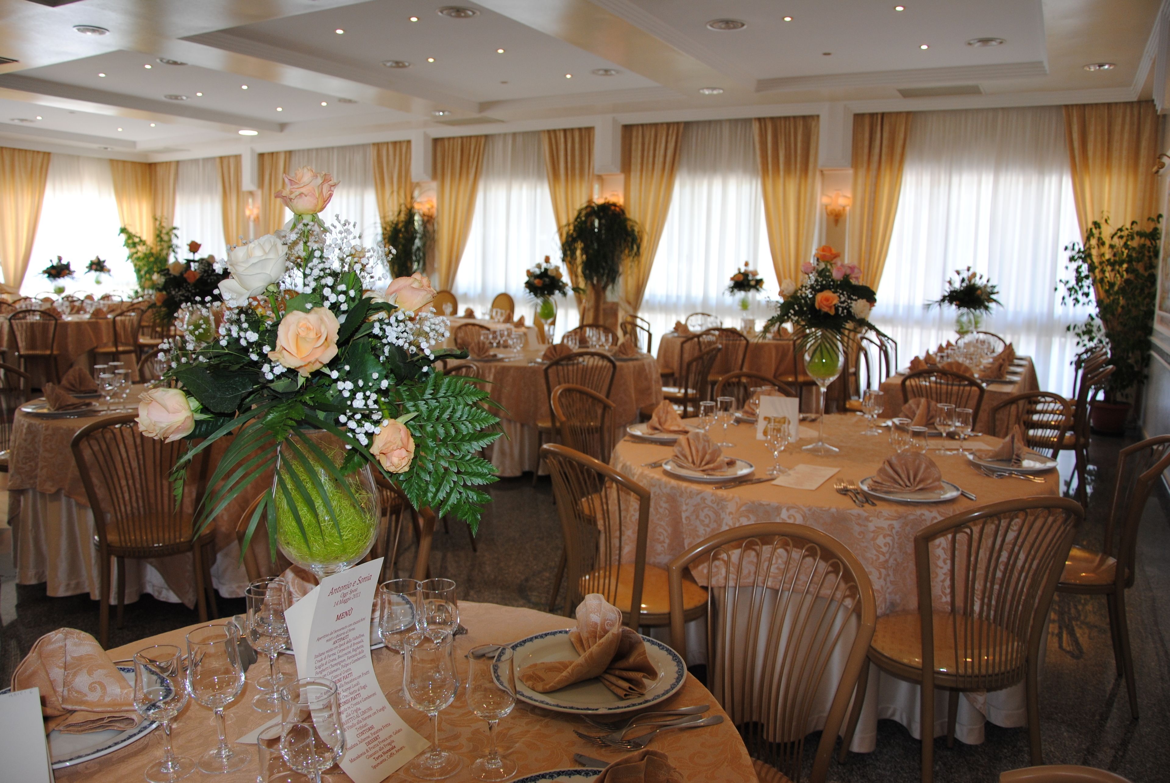 A banquet hall with round tables set for dining, each with a menu stand, wine glasses, and floral centerpieces.