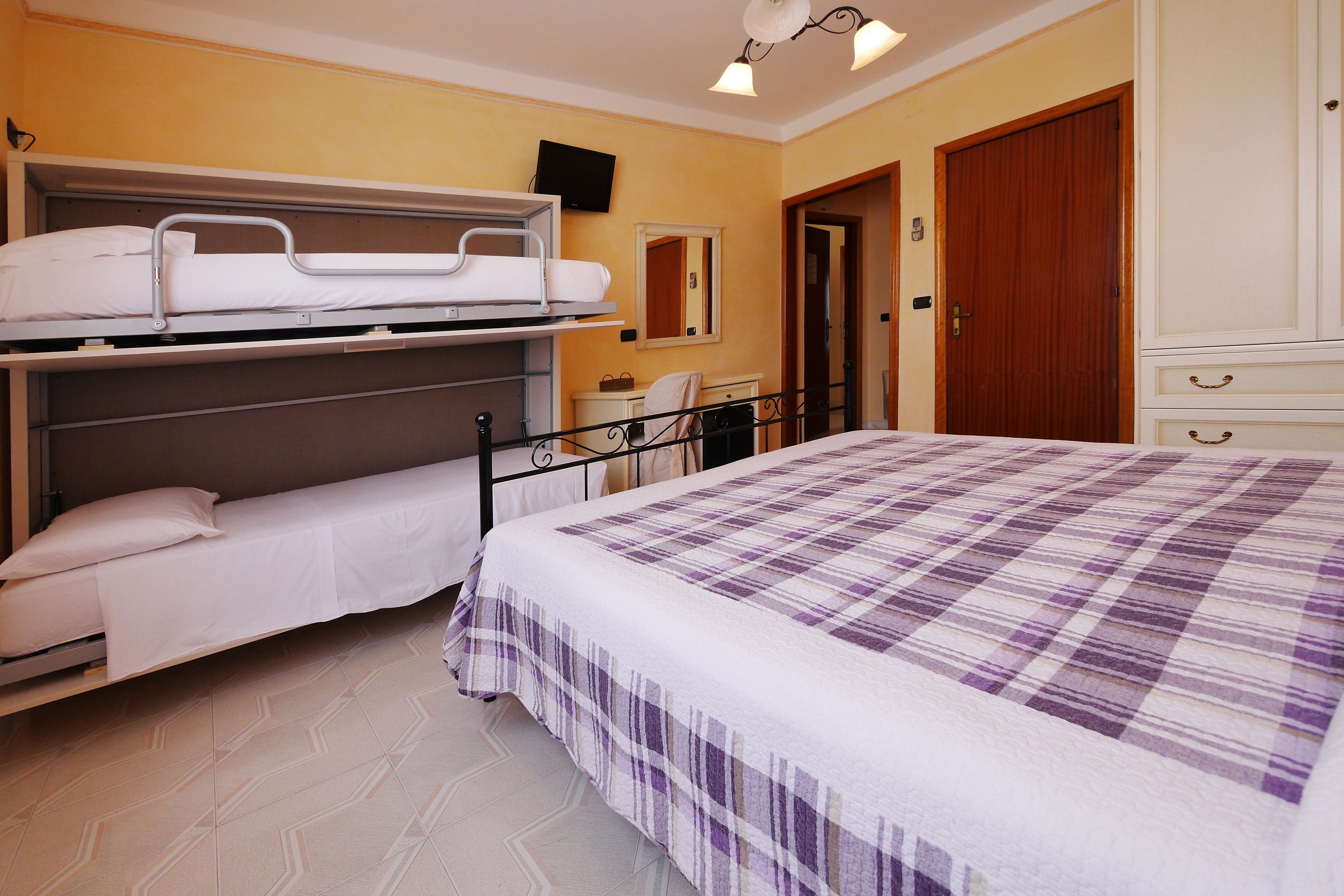 A hotel room with a bunk bed, a single bed, a bedside table, a mirror, and a wardrobe.