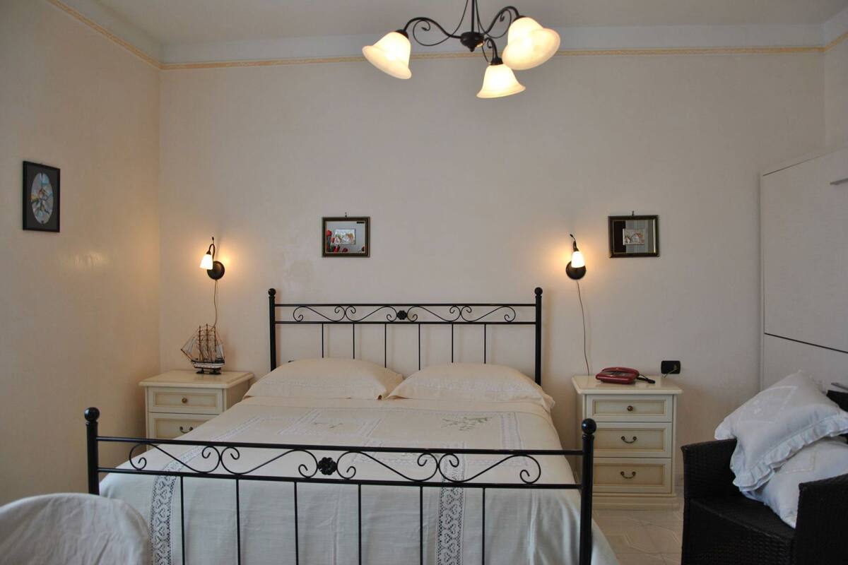Une chambre à coucher avec un lit en métal, deux tables de chevet, une commode, une chaise en osier et un lustre.