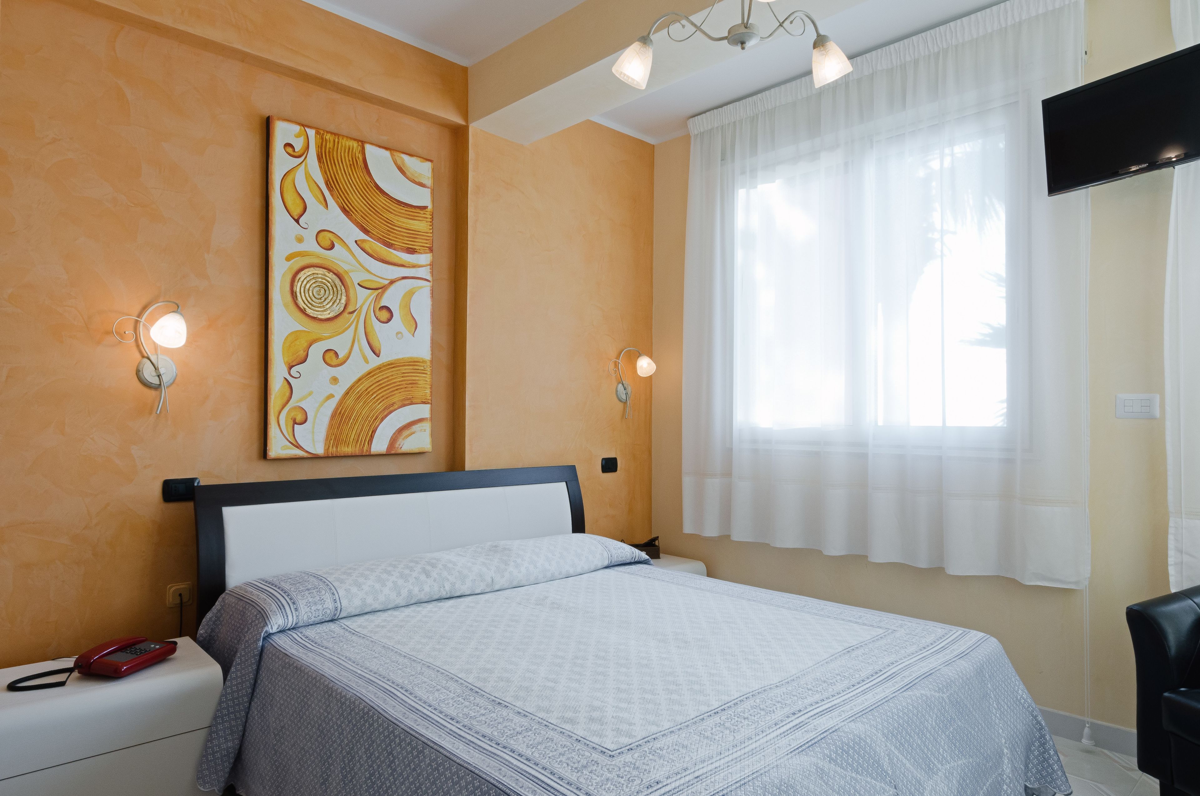 A hotel room with a bed, a painting on the wall, two wall-mounted lights, a TV, and a window with curtains.