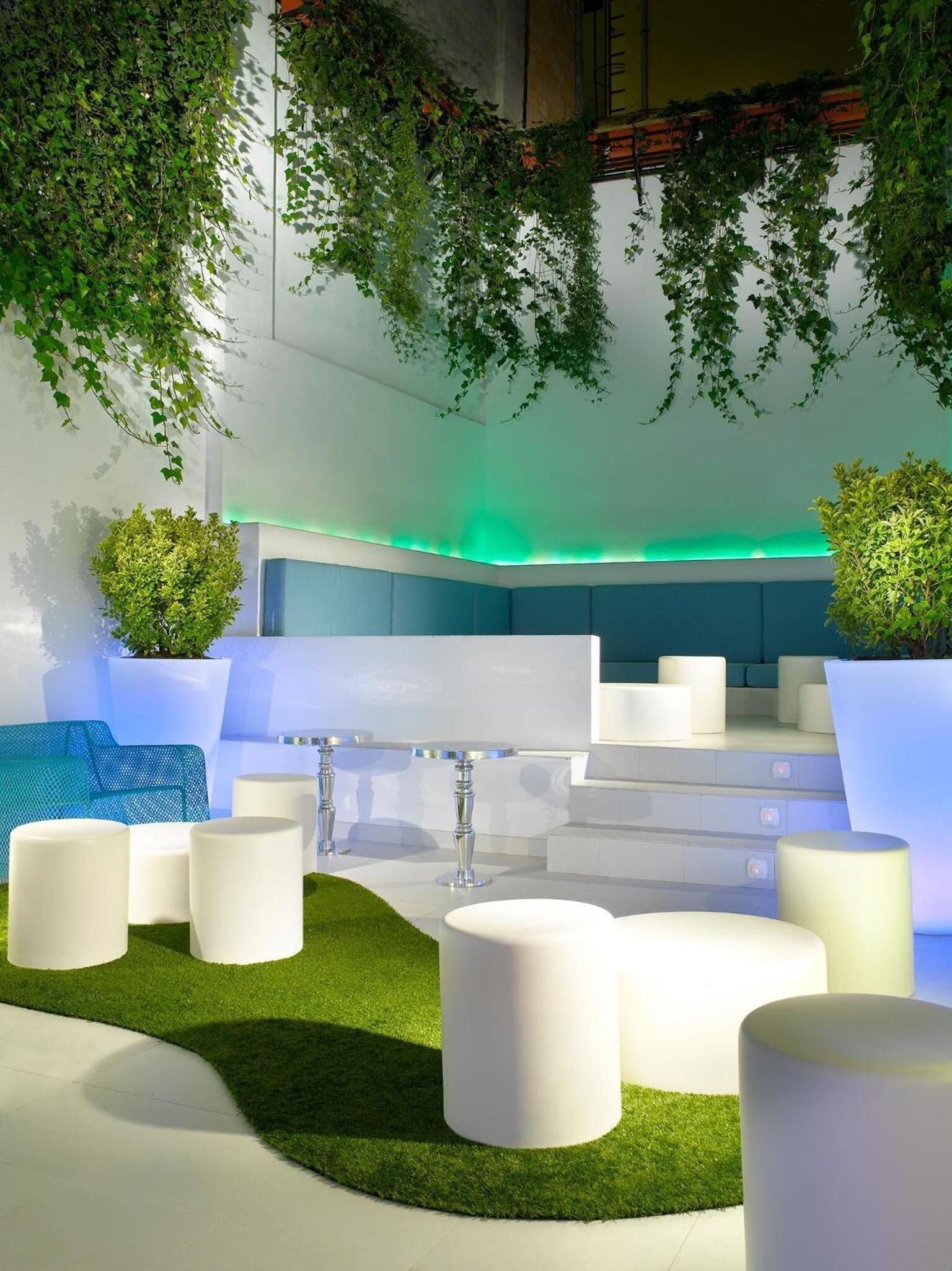 A modern outdoor seating area with white cylindrical stools, a blue sofa, and green plants.