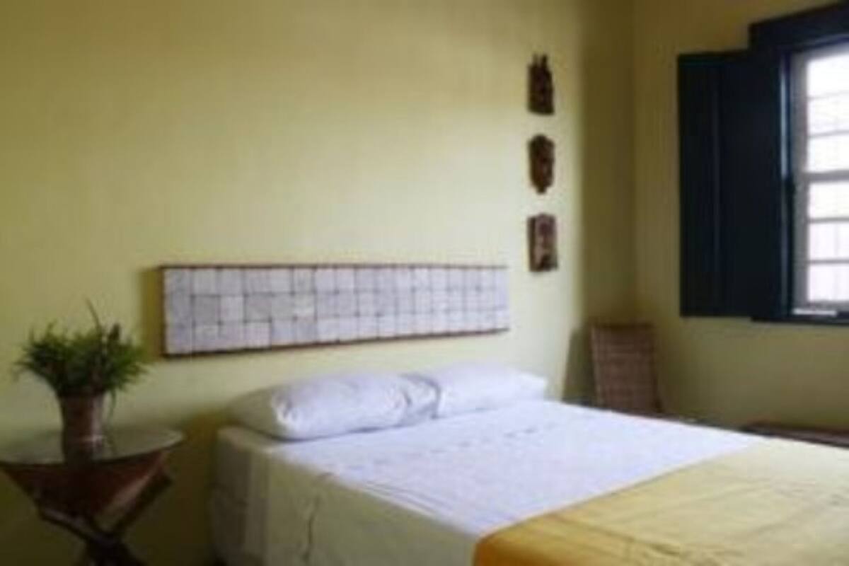 A bedroom with a bed, a bedside table with a plant, a window with blinds, and a wall-mounted artwork.