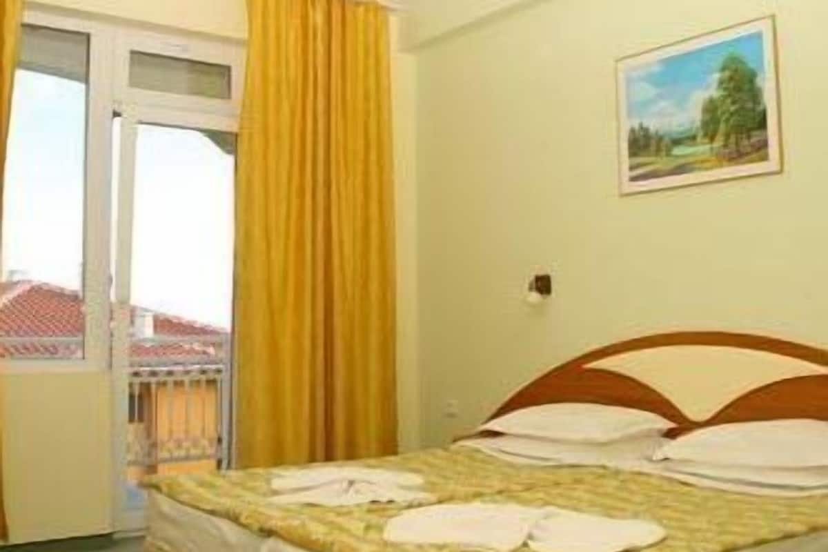A hotel room with a bed, a window with yellow curtains, and a painting on the wall.