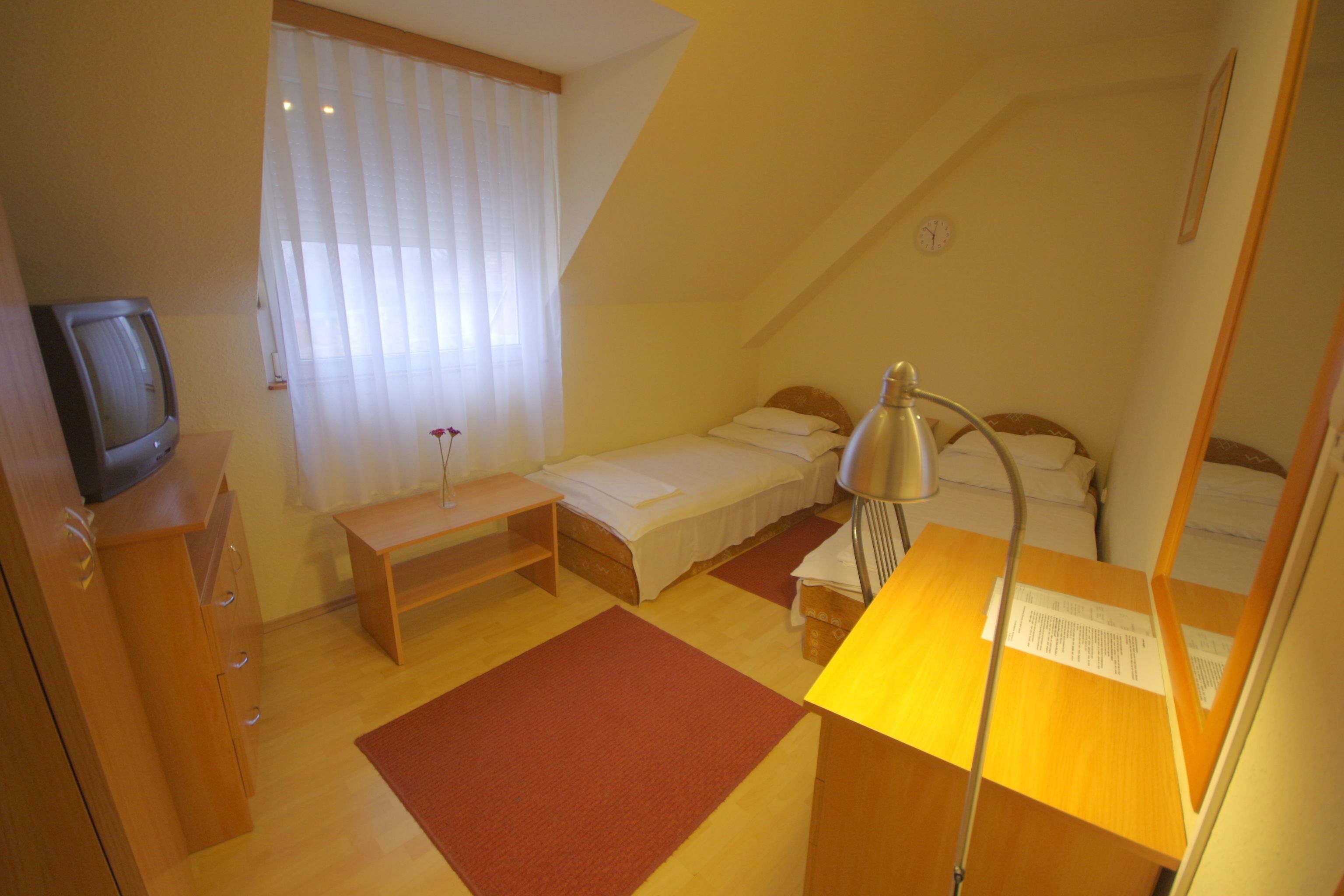 A small, well-lit bedroom with a single bed, a desk with a lamp, a television, and a window with curtains.