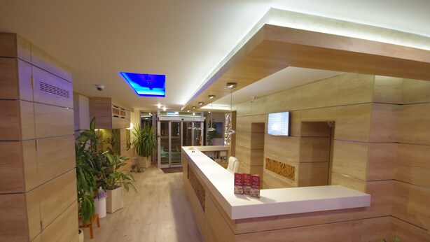A modern reception area with a wooden reception desk, potted plants, and a glass elevator. - Hotel Vadászkürt Superior (Szekesfehervar)