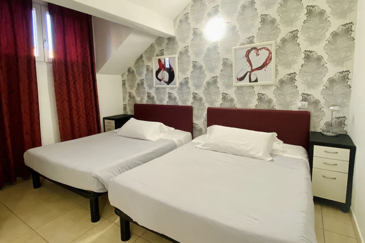 Una habitación con dos camas, una cortina roja, un papel tapiz con motivos de hojas y dos cuadros enmarcados.