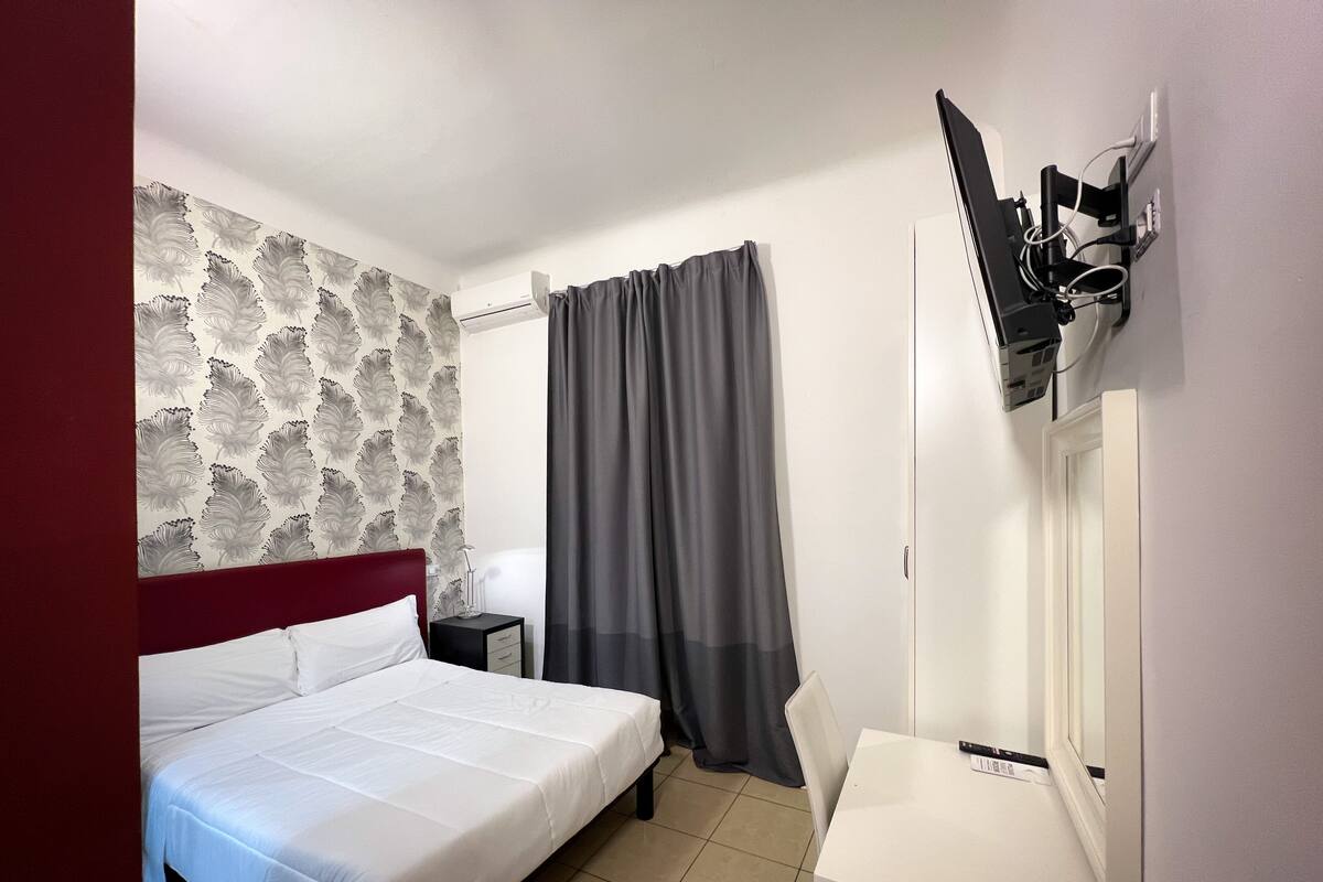 Habitación de hotel con cama, mesita de noche, televisor montado en la pared y ventana con cortinas.