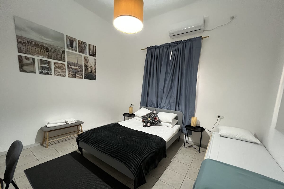 Ein Schlafzimmer mit einem Bett, Nachttischen, einem Sessel, einem kleinen Tisch und einer Wand-Klimaanlage.