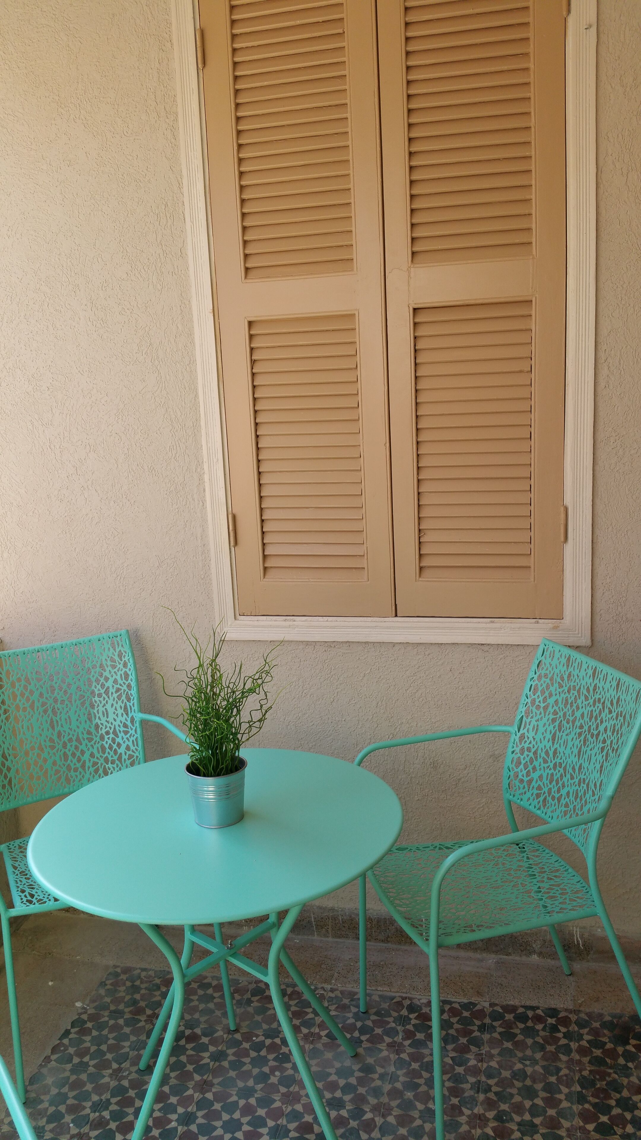 A small outdoor seating area with a round table and two chairs, a potted plant, and a window with shutters.