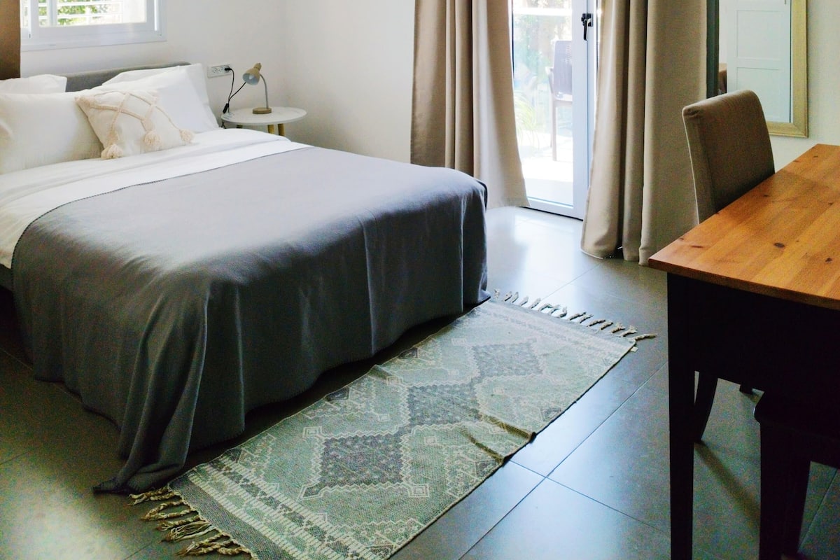 Ein Schlafzimmer mit Bett, Schreibtisch, Stuhl, einer Pendelleuchte und einem Fenster mit Vorhängen.