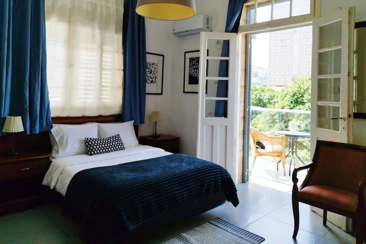 Ein Schlafzimmer mit Bett, Nachttisch, Lampe, einem Fenster mit Vorhängen und Blick ins Freie.