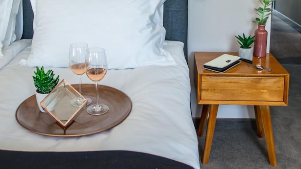 Una cama bien hecha con ropa blanca, una mesita de noche con un libro, un florero y una planta pequeña. Hay dos copas de vino sobre una bandeja.
