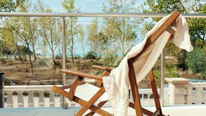 Una silla de madera con cojín blanco, ubicada en un balcón con vistas a árboles y una masa de agua.
