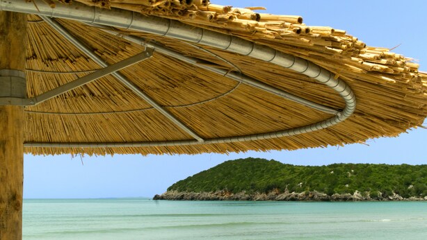 Una sombrilla de playa hecha con materiales naturales, una estructura de madera y vistas al mar y una colina verde.