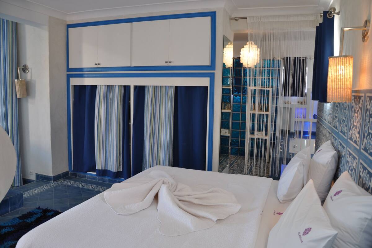 A hotel room with a bed, curtains, a wardrobe, and a bathroom visible through a glass door.