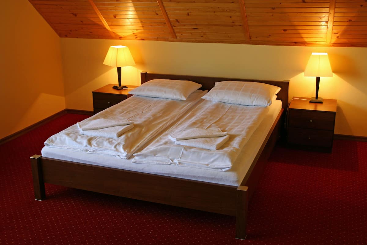 A neatly made bed with white linens in a room with wooden ceiling beams and red carpet flooring.