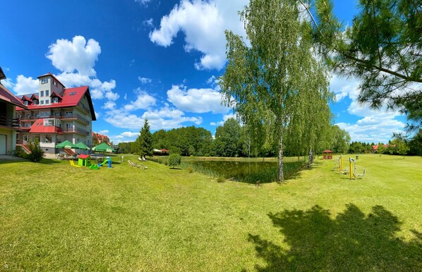 A grassy area with a pond, trees, and a building with a red roof. - Hotel Mazury (Gizycko)