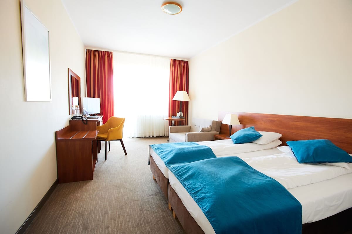 A hotel room with a desk, two beds, a chair, and a lamp.