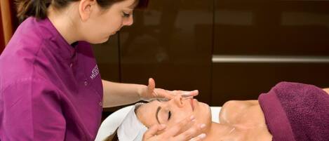 A person receiving a facial treatment in a room with a dark brown cabinet and a marble countertop.