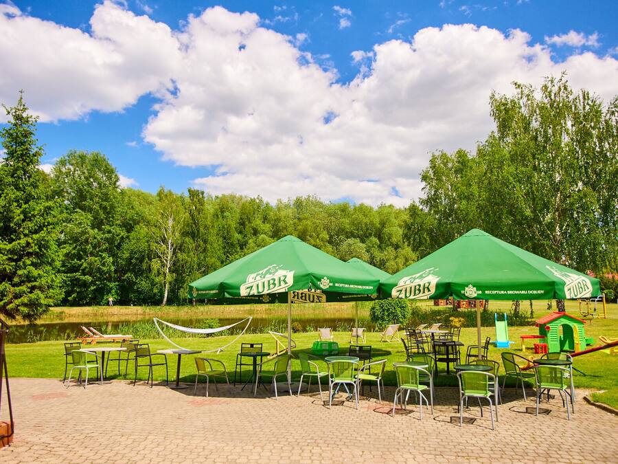 An outdoor seating area with green tables and chairs under green umbrellas, a pond, and trees in the background.