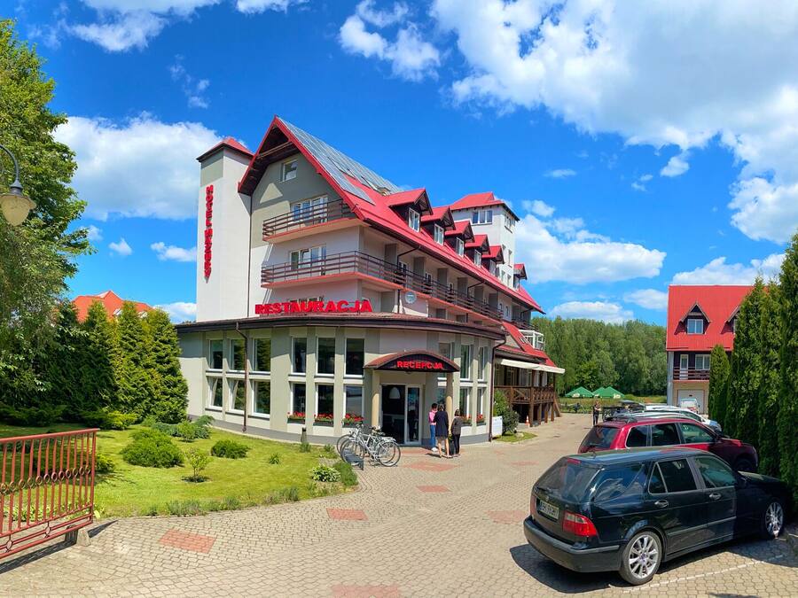 A hotel with a red roof and multiple balconies, a reception area, and a paved driveway with parked cars.