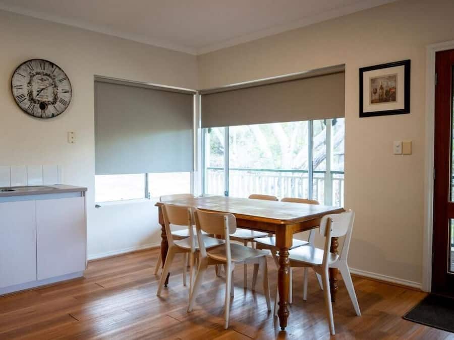 A dining area with a wooden table and chairs, a clock on the wall, and a window with a view of trees.
