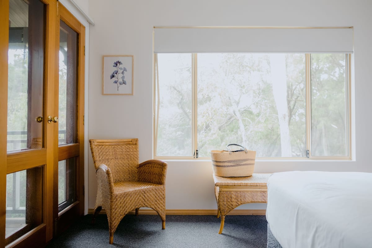 A room with a large window, a wicker chair, a bench with a woven basket, and a bed.