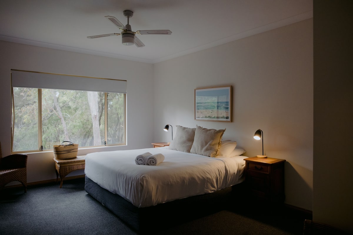 A bedroom with a large bed, a ceiling fan, a window with a view of trees, a bedside table with a lamp, and a framed picture on the wall.