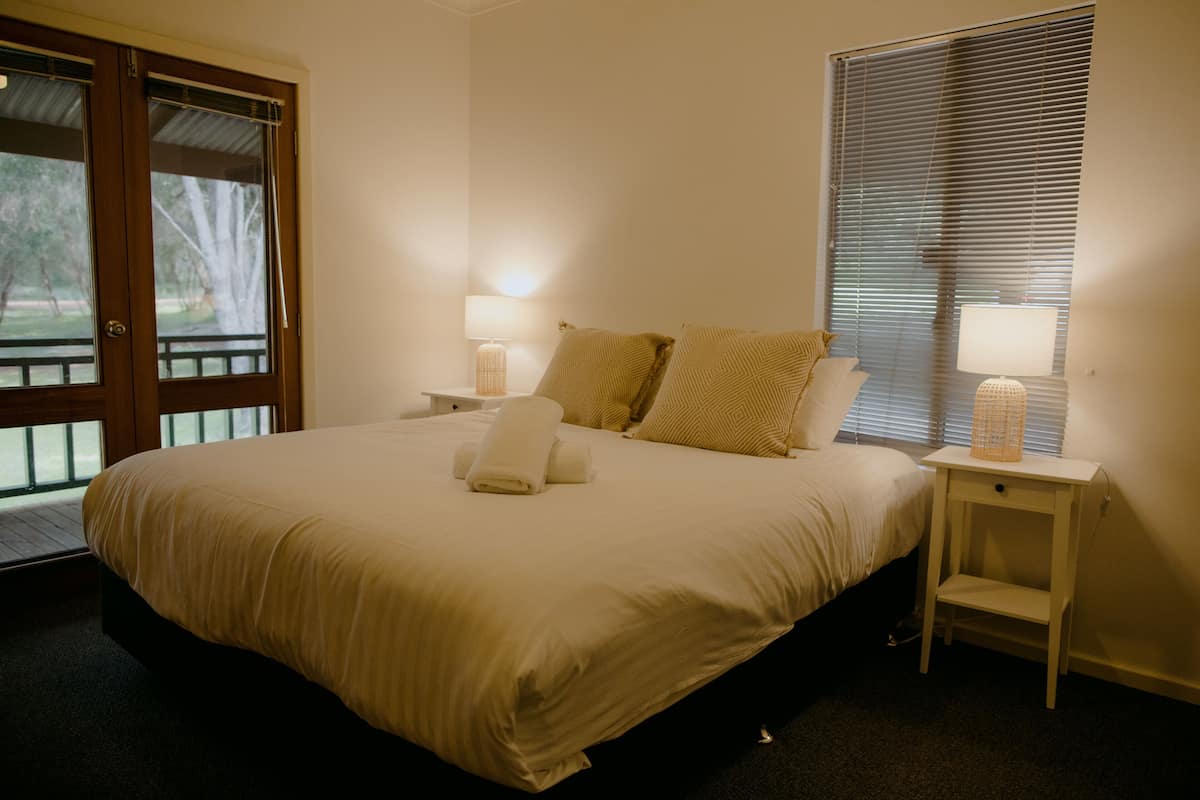 A neatly made bed with pillows and a folded towel, a bedside table with a lamp, and a window with blinds.