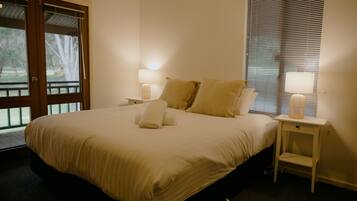 A neatly made bed with pillows and a folded towel, a bedside table with a lamp, and a window with blinds.