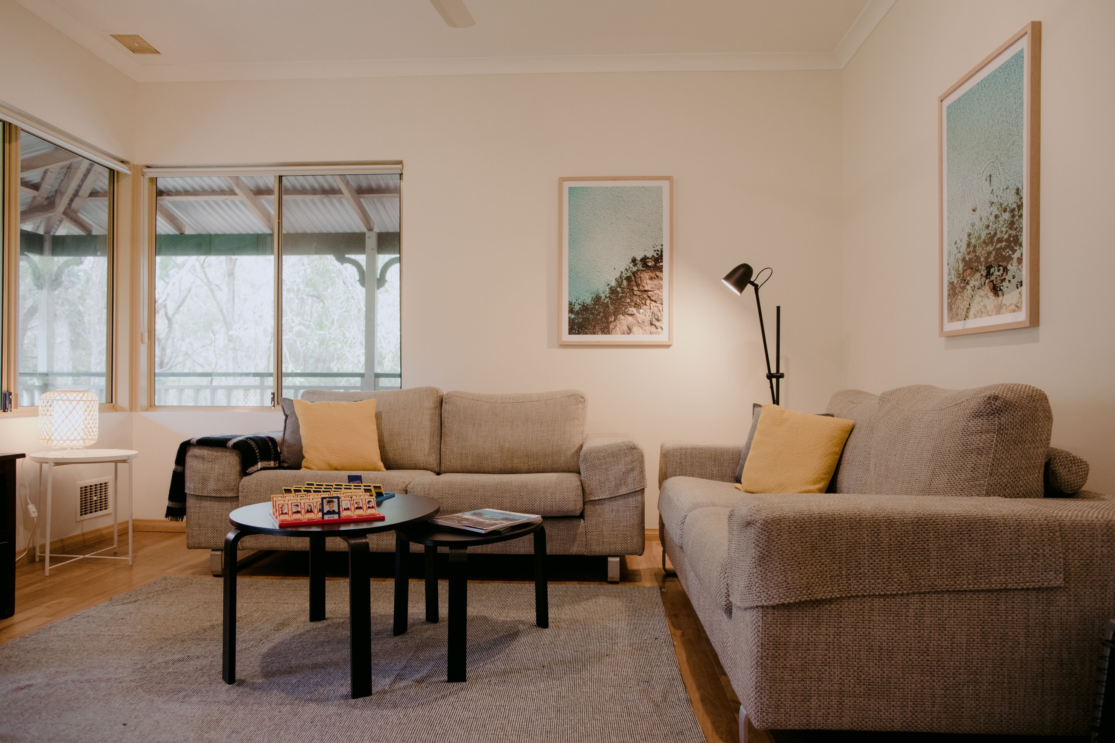Ein Wohnzimmer mit zwei Sofas, einem Couchtisch und einer Lampe. An den Wänden hängen Bilder und ein Fenster bietet Blick auf Bäume.