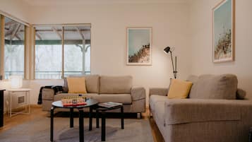 A living room with two sofas, a coffee table, and a lamp. There are framed pictures on the walls and a window with a view of trees.