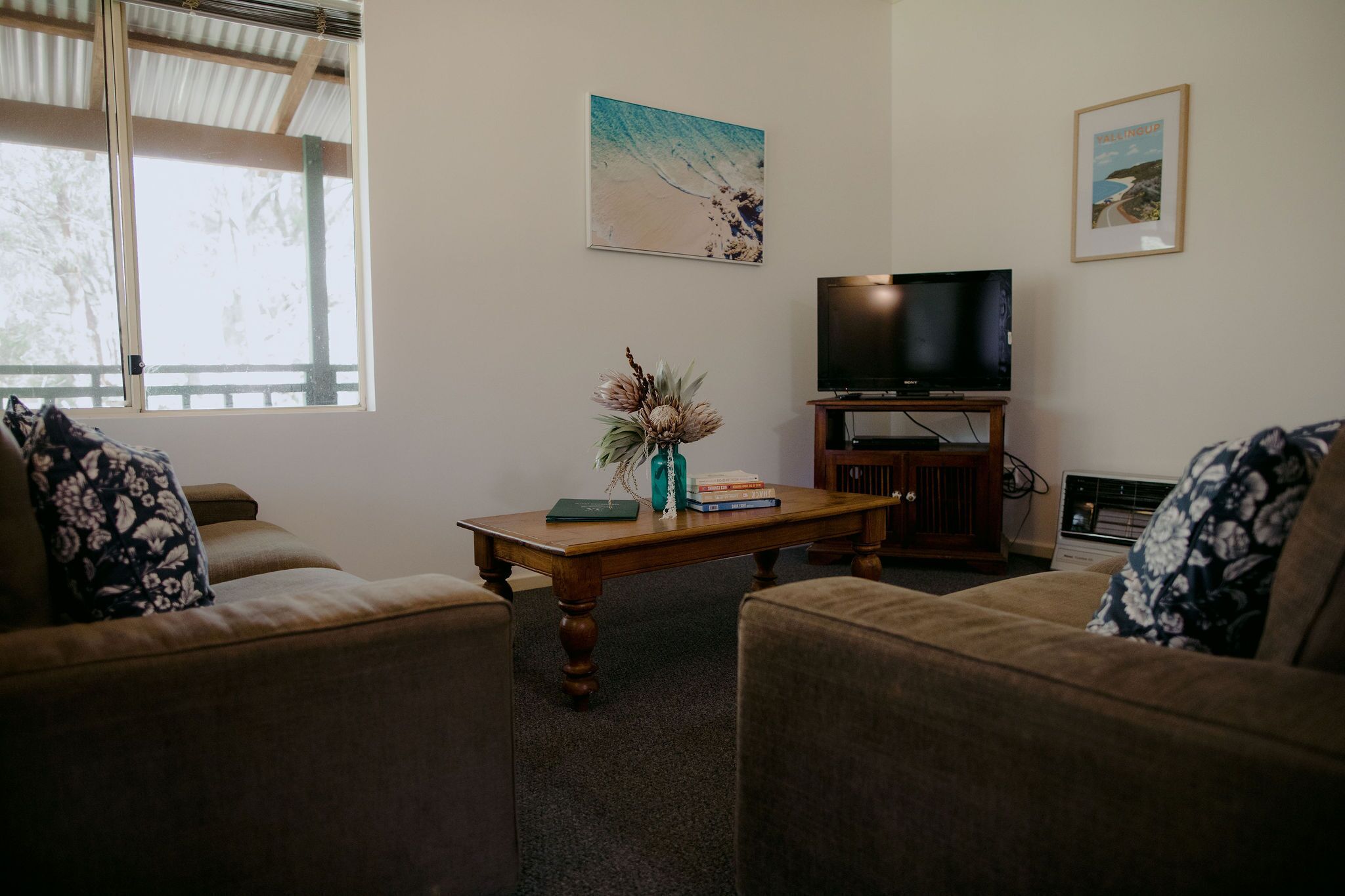 Una sala de estar con un sofá, una mesa de centro con libros y un jarrón, un televisor sobre un mueble bajo, y dos cuadros en la pared.