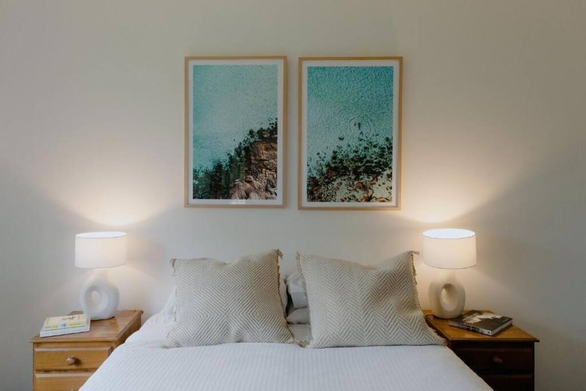A neatly made bed with two pillows, a bedside table with a lamp, and two framed pictures on the wall.