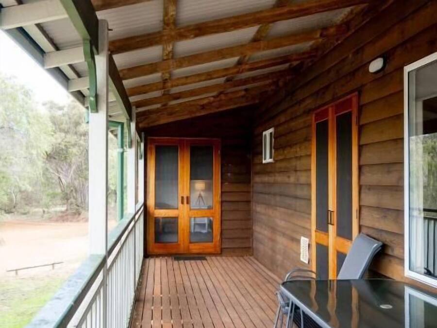 Una terraza de madera con una mesa y sillas de vidrio, una puerta con panel de vidrio y vistas al exterior.