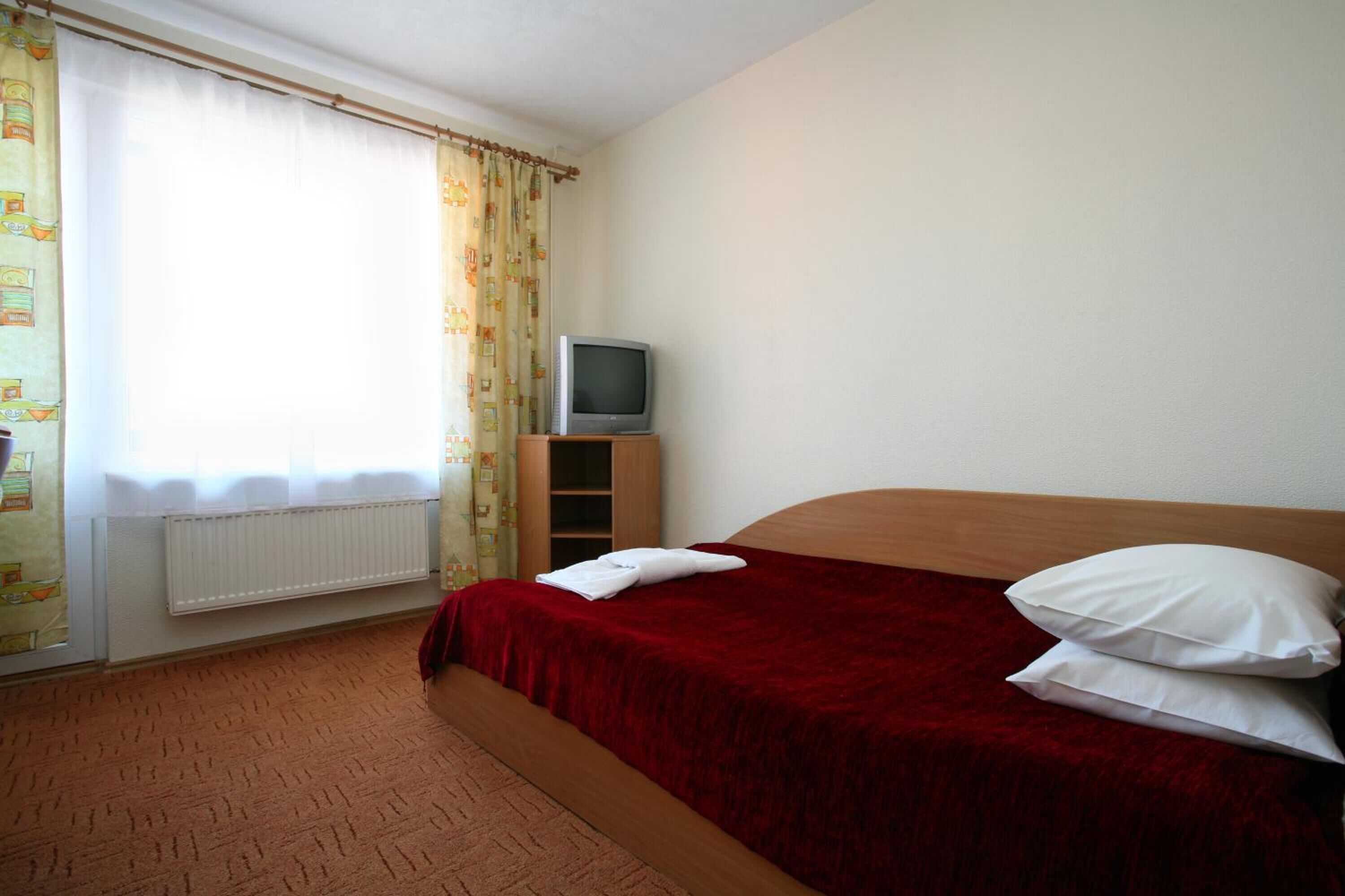 Quarto de hotel com cama, televisão sobre suporte de madeira, radiador e cortinas.