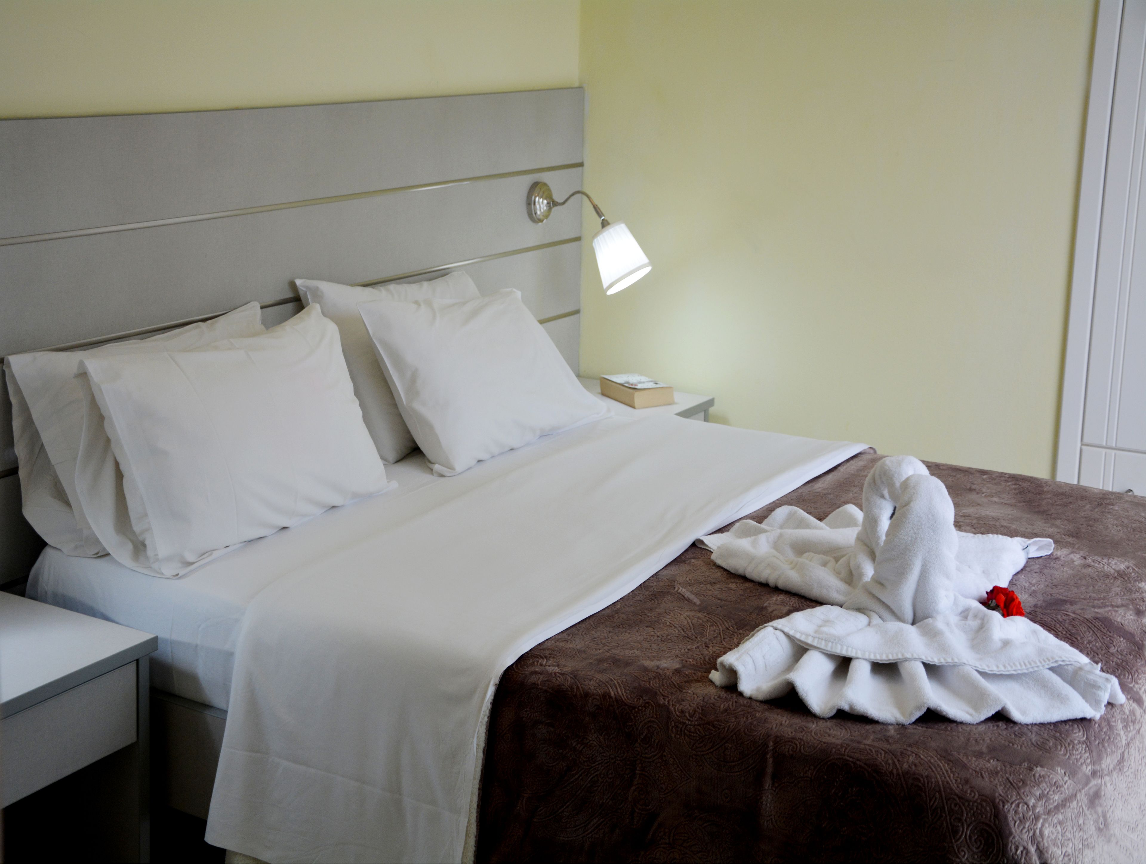 Una cama bien hecha con sábanas blancas y una manta marrón, una mesita de noche con una lámpara, y un mueble auxiliar con una caja.