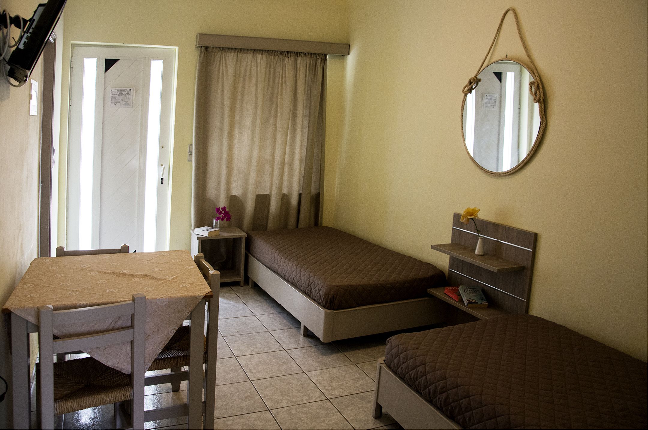 A small, well-lit room with a bed, a desk, and a chair. There is a mirror on the wall and a window with curtains.