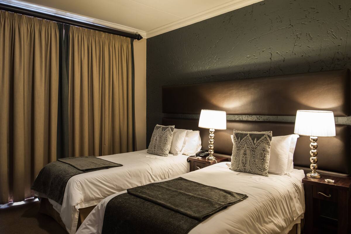 Habitación de hotel con dos camas, un ventanal grande con cortinas, cabecero, lámparas de noche y mesita de noche con teléfono.