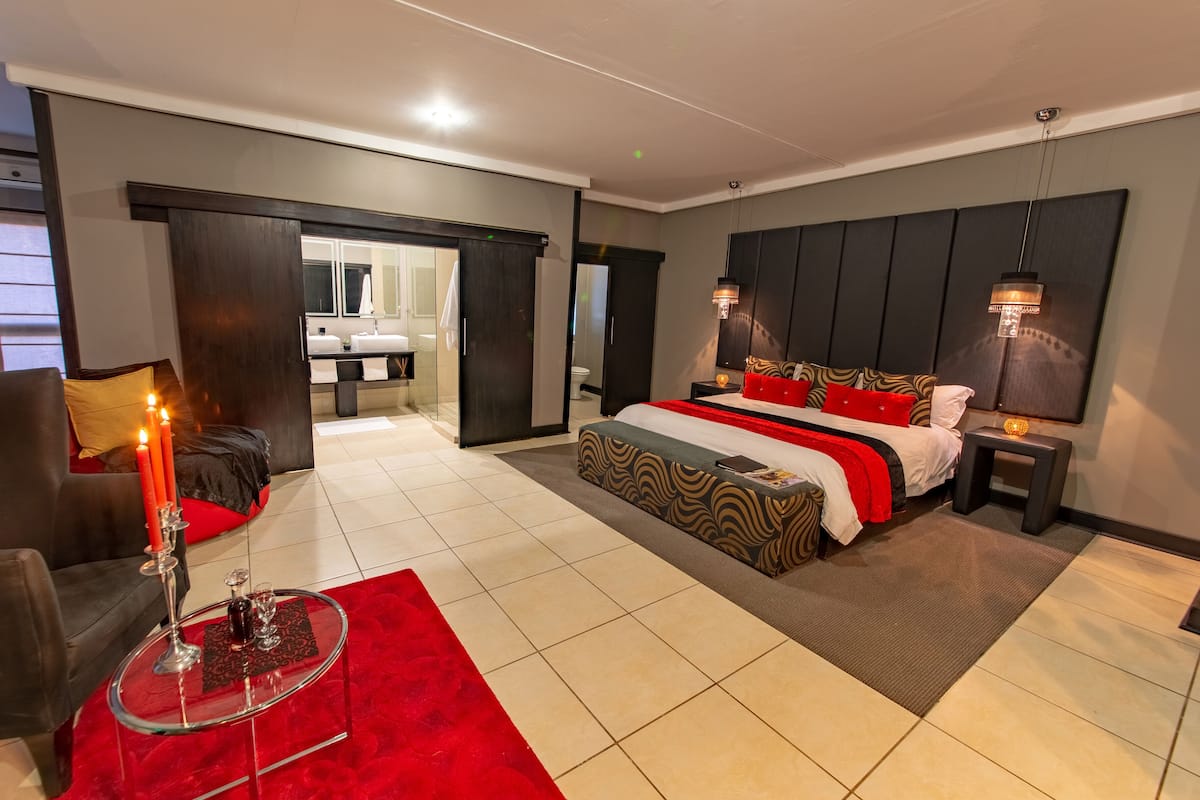 Une chambre d’hôtel moderne avec un grand lit, un coin salon agrémenté d’un tapis rouge, une table basse en verre et une salle de bain visible à travers une porte ouverte.