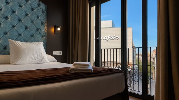 Une chambre d’hôtel avec une tête de lit capitonnée, un lit recouvert de draps blancs, une serviette pliée et une vue sur la ville à travers de grandes fenêtres.