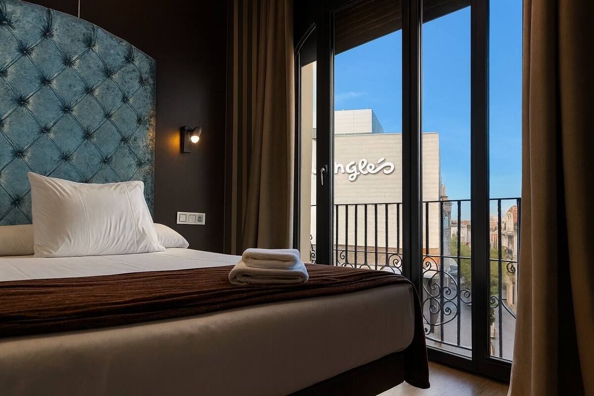 A hotel room with a tufted headboard, a bed with white linens, a folded towel, and a view of the city through large windows.