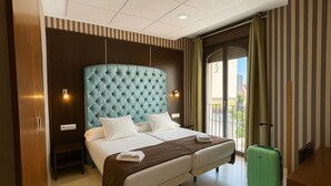 Une chambre d’hôtel avec un grand lit, une valise verte et une vue sur la ville par la fenêtre.