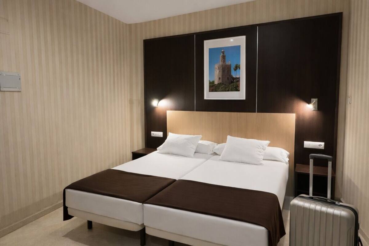 A hotel room with a double bed, a bedside table, a lamp, a picture on the wall, and a suitcase.