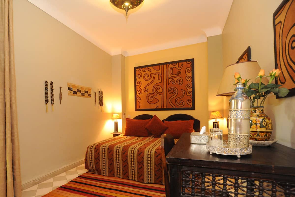 A hotel room with a bed, a nightstand, a table with a vase and a bottle, and a wall with decorative patterns.